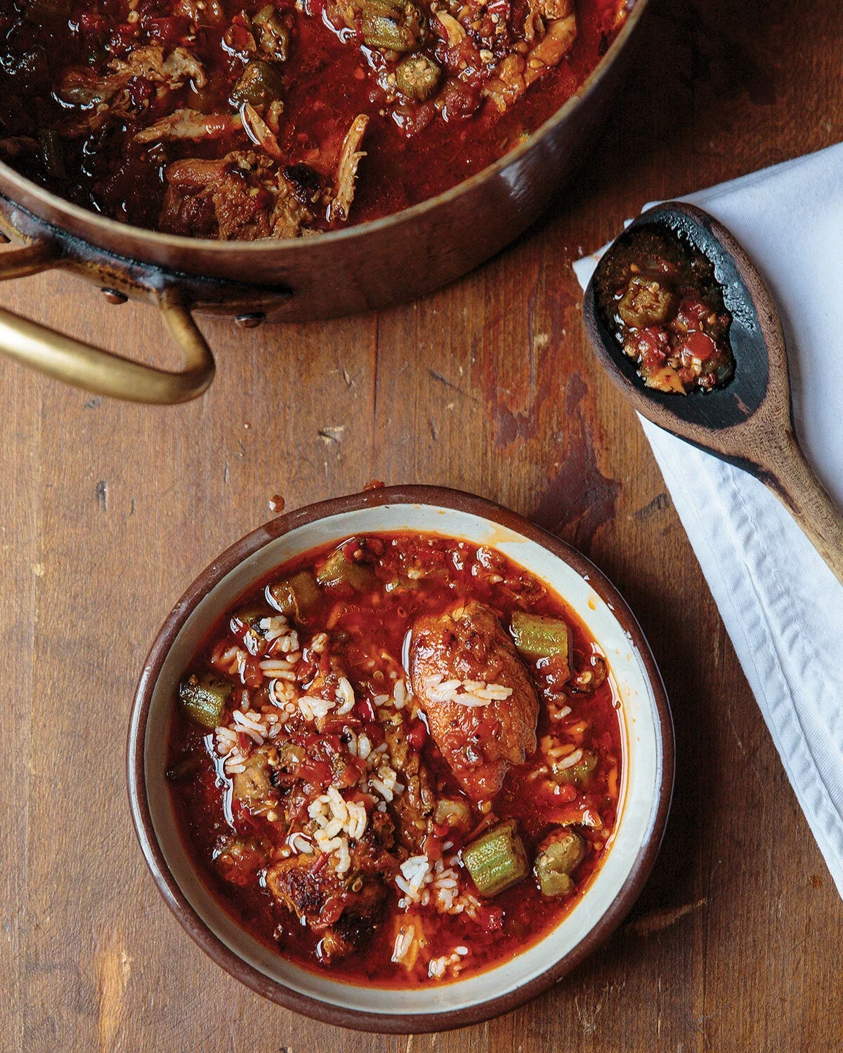 Bowl of tomato-red okra gumbo with rice and pieces of meat on a wooden table, with a pot of gumbo and a wooden spoon nearby.