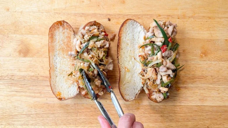 Using tongs to pile cooked chicken, peppers, and onions on hoagie buns