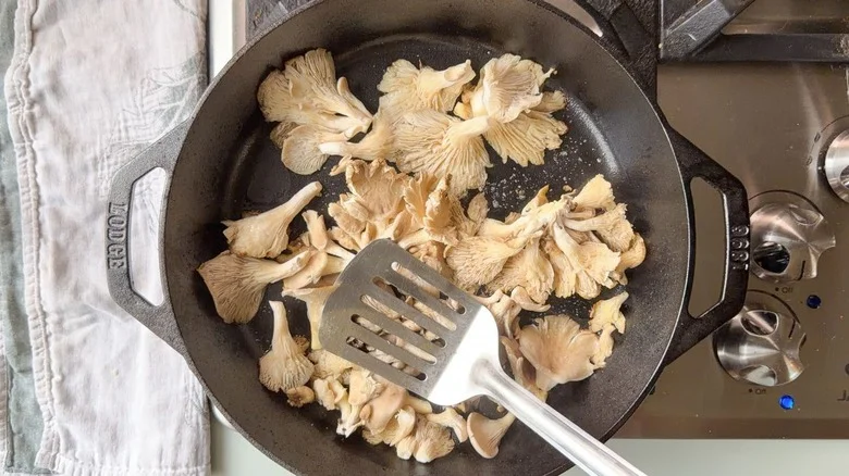 Pressing down on oyster mushrooms in cast iron skillet with metal spatula on stovetop
