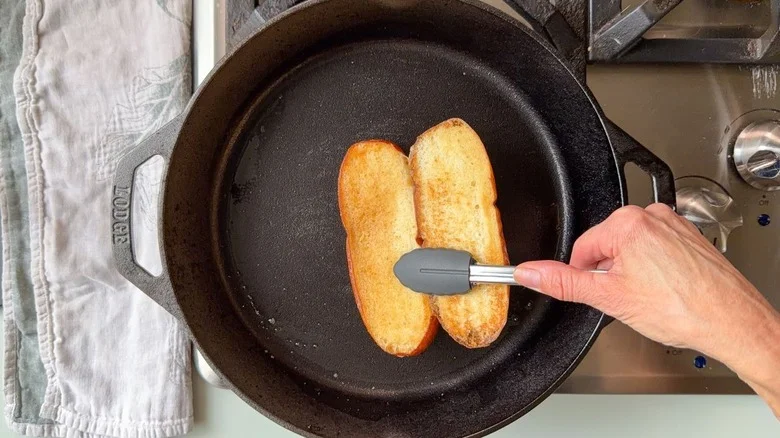 Using tongs to flip a toasted pretzel hoagie bun in cast iron skillet on stovetop