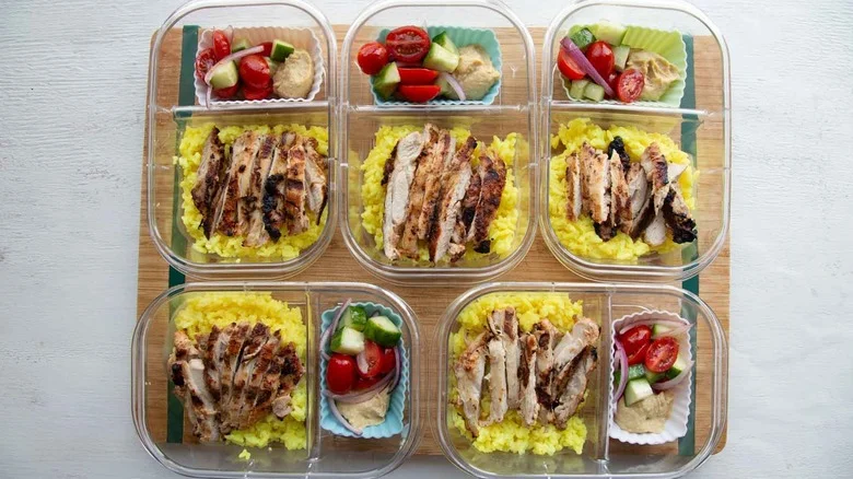 five meal prep containers with chicken, rice, and silicone muffin tin with hummus and tomato cucumber salad