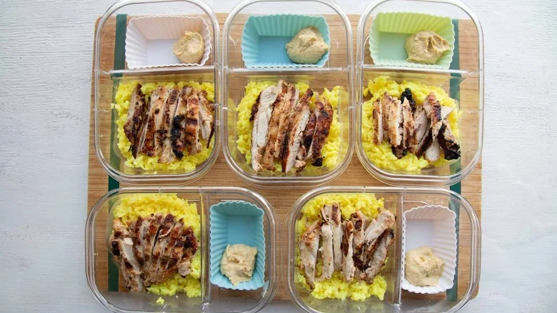 five meal prep containers with chicken, rice, and silicone muffin tin with hummus