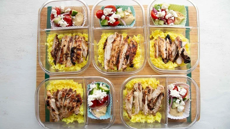 five meal prep containers with chicken, rice, and silicone muffin tin with hummus and tomato cucumber feta salad
