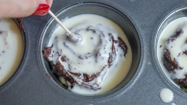 Hand using toothpick to swirl cheesecake batter into brownie batter in muffin tin