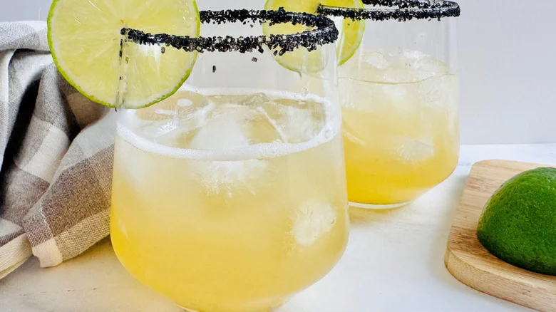 two drinks in glasses with black salt rims and lime garnishes in front of a crumpled checked cloth napkin