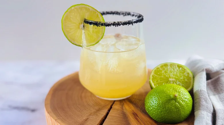 drink in a glass with a black salt rim and a lime garnish sitting on a circular chunk of wood next to a crumpled checked cloth napkin