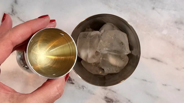 hand holding a shot glass of liquid above a metal tumbler