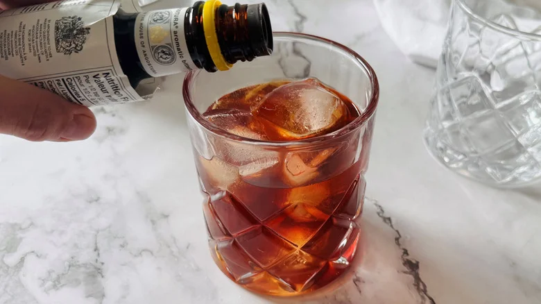 hand holding bitters bottle above a drinking glass filled with ice and brown liquid
