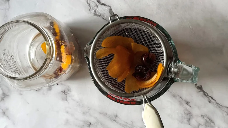 orange peel and cherries in a strainer above a jar