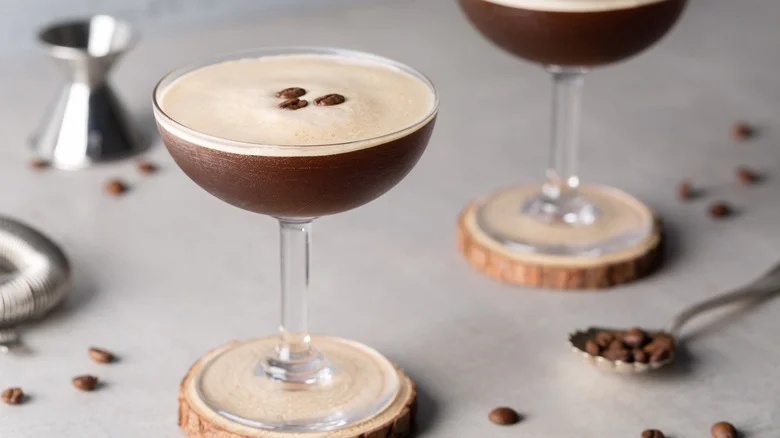 A frothy espresso martini in a round martini glass on a round wood coaster.