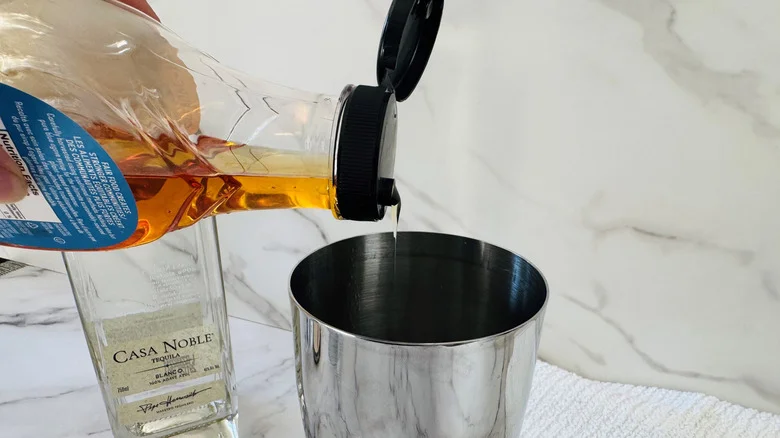 hand pouring brown liquid from a bottle into a cocktail shaker